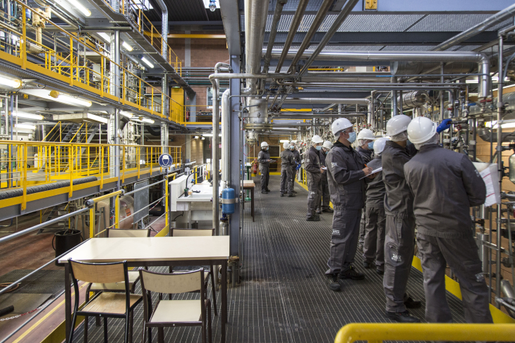 Une dizaine de personnes regardent les installations d'une usine équipés de leurs EPI (combinaison, casques, masques, lunettes, gants) et prennent des notes sur ce qu'ils observent.
