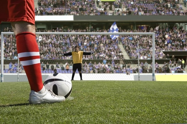 Sur un terrain de foot, un joueur en rouge a le ballon devant son pied et le gardien en jaune devant les cages tend les bras à l'horizontal prêt à réceptionner le ballon.